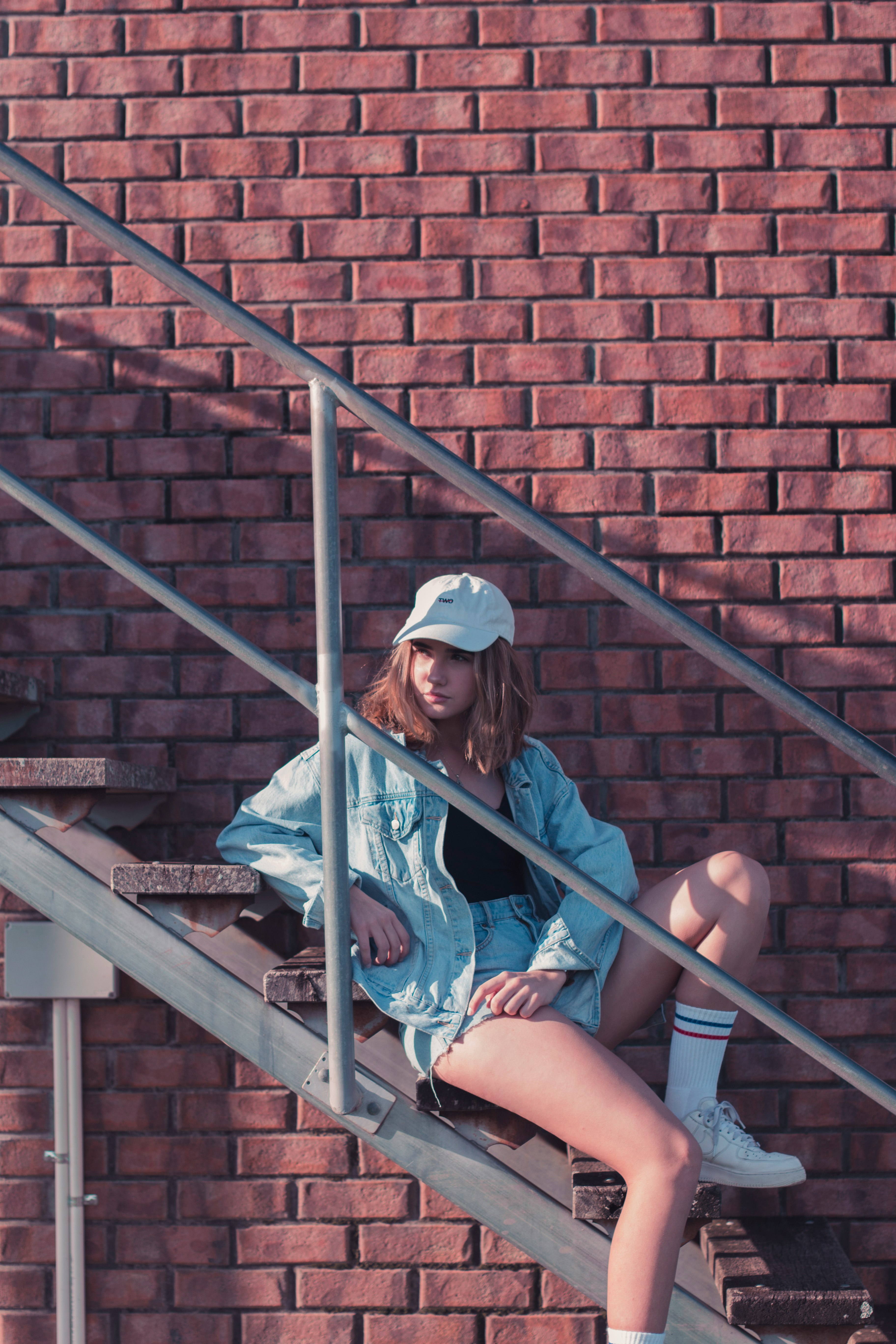 woman wearing gray denim jacket sitting on brown wooden stair near brown concrete wall during daytime
